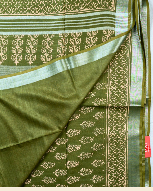 An olive green pure linen cotton saree with beige block prints and a silver border, shown folded on a light background with a brand logo banner at the bottom.